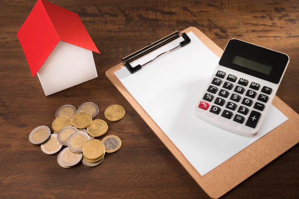 A desk with a calculator, coins, and a small house model with a red ceiling.