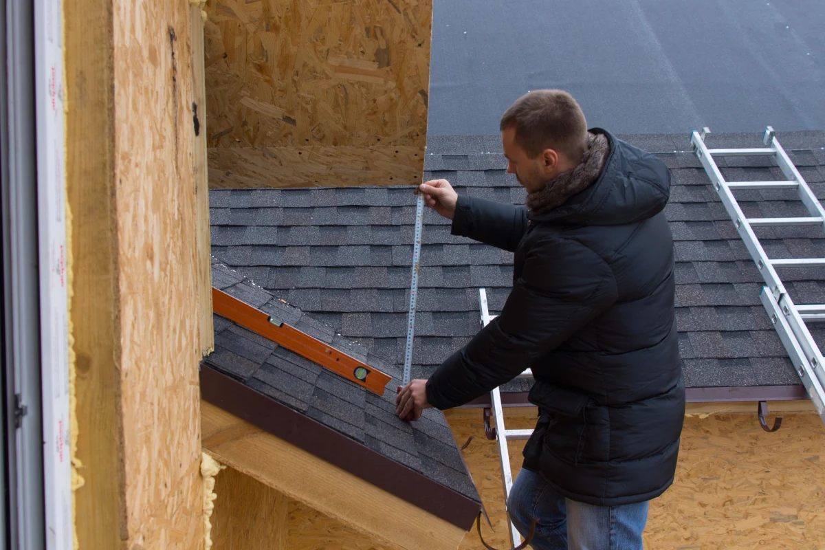 A roofer measures a roof for shingles on a new house using a tape measure while standing on a ladder during the roof installation.