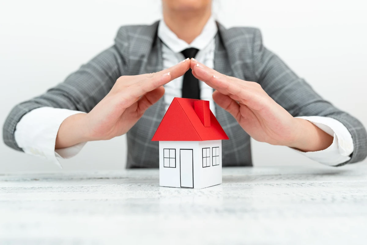 Woman in suit and tie holding up paper house model, hand positioned like a roof.