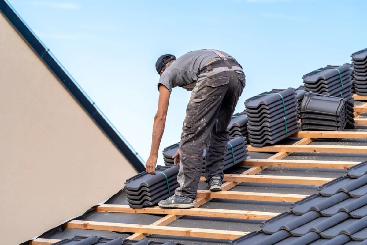 A man is replacing a black tiled roof, standing on the roof while working diligently on the project.